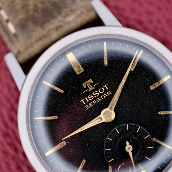 A close-up of the Tissot Seastar Black Gilt Dial Sub Second showcases its black dial, gold hour markers and hands, and a second subdial. The silver-toned case is paired with a worn brown leather strap, set against a textured red surface.