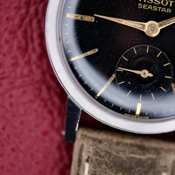 A close-up showcases the Tissot Seastar Black Gilt Dial Sub Second wristwatch, featuring a black face with gold hands and a small second hand sub-dial, encased in silver and paired with a textured brown strap, set against a maroon textured background.