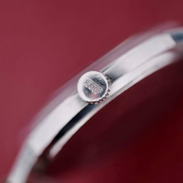 A close-up shows the silver watch crown engraved with Tissot from the Tissot Seastar Black Gilt Dial Sub Second. The background is muted red, while the focus highlights the crowns metallic texture.