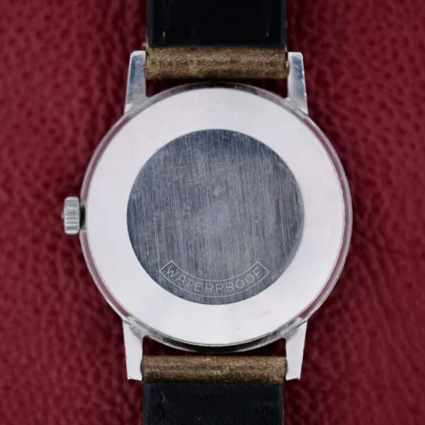 The image features the back of a Tissot Seastar Black Gilt Dial Sub Second wristwatch, showcasing a round metal casing with WATERPROOF engraved. It has a black leather strap against a textured red background.