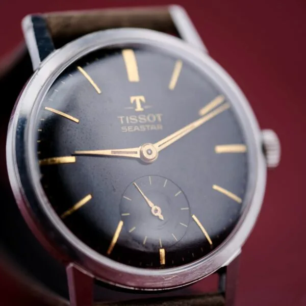 Close-up of a Tissot Seastar Black Gilt Dial Sub Second wristwatch, featuring gold-tone hands and a sub-dial above 6 oclock in a round silver casing, set against a blurred dark red background.