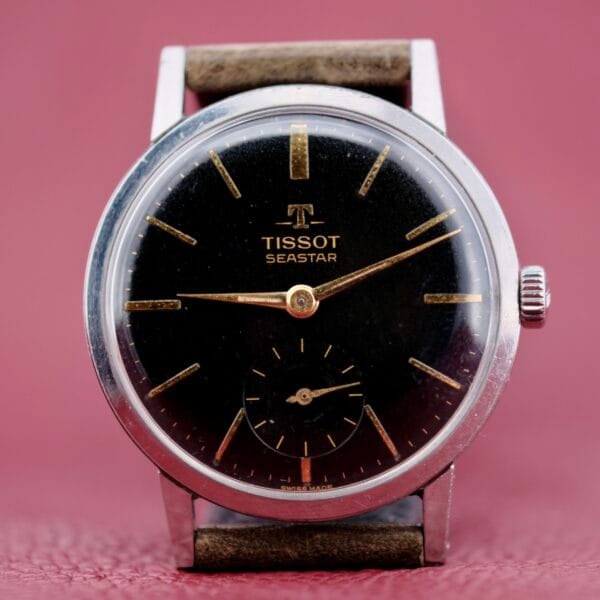 The Tissot Seastar Black Gilt Dial Sub Second is a vintage wristwatch featuring a black dial, leather strap, silver hour markers, slender hands, and a small second subdial at six oclock against a pink textured background.