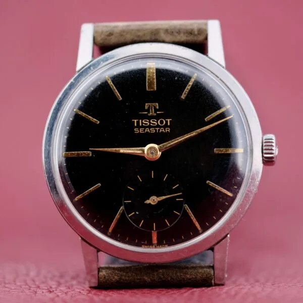 The Tissot Seastar Black Gilt Dial Sub Second is a vintage wristwatch featuring a black dial, leather strap, silver hour markers, slender hands, and a small second subdial at six oclock against a pink textured background.