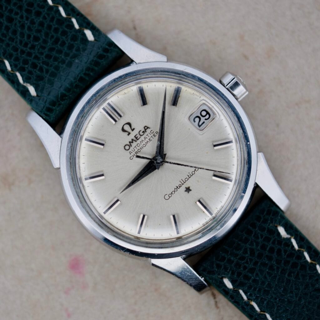 The Omega Constellation Chronometer 14939-1 Flat Dial is a silver wristwatch with a black strap, date display, and black hour markers on a beige dial.