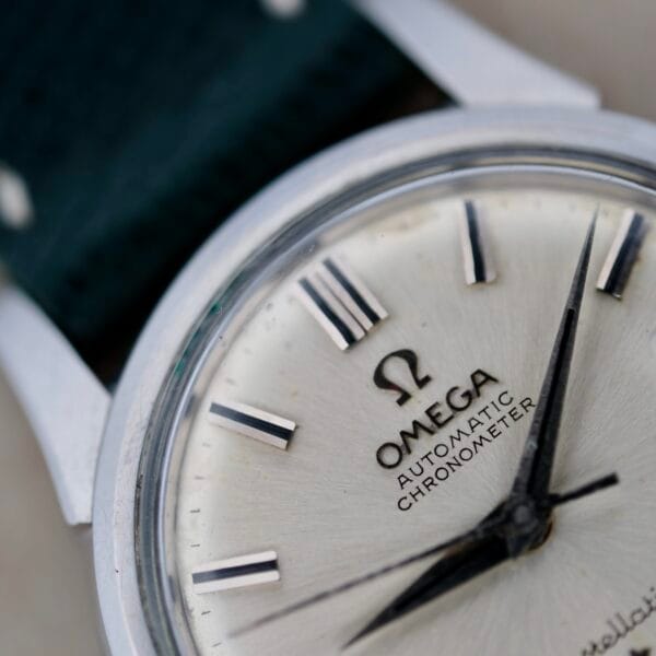 Close-up of a vintage Omega wristwatch with a silver face and black hour markers. The brand logo and Automatic Chronometer grace the dial. This exquisite piece, from the renowned collection of vintage watches, features a dark strap and stainless steel case.