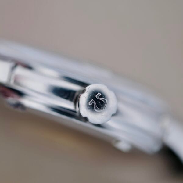 Close-up of a silver watch crown with an engraved S, echoing the elegance of the Omega Constellation Chronometer 14939-1 Flat Dial. The case and part of the bracelet emerge against a blurred backdrop, highlighting the crowns exquisite details.