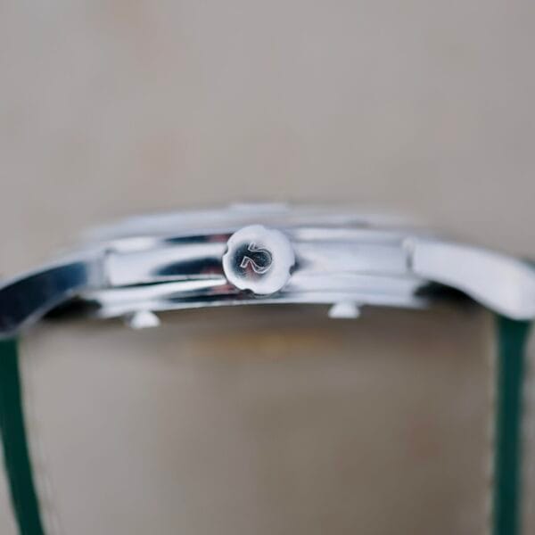 Close-up of a sleek silver watch case viewed from the side, highlighting a crown embossed with R. The Omega Constellation Chronometer 14939-1 Flat Dial exudes elegance with its green strap against a blurred background that enhances its metallic allure.