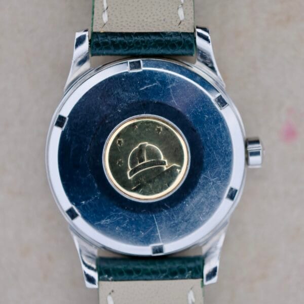 The image displays the back of an Omega Constellation Chronometer 14939-1 Flat Dial, featuring a circular emblem with a hard hat design. The vintage watch has a metallic case showing scuffs and scratches and is complemented by a green and beige leather strap, highlighting its timeless allure.