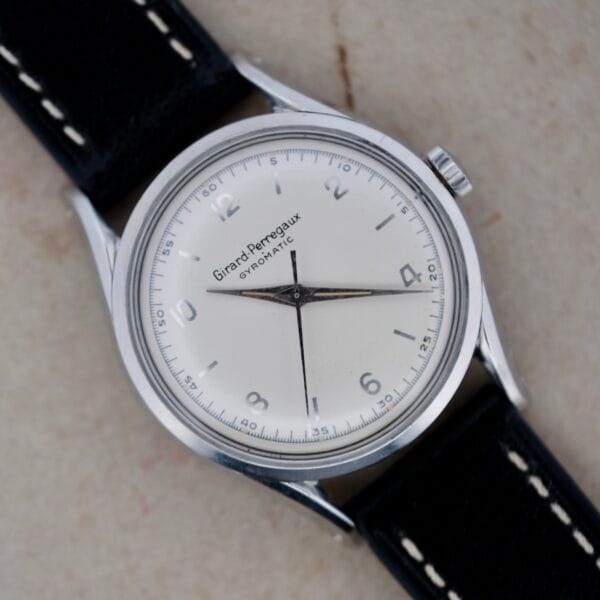 The vintage Girard Perregaux Gyromatic 6169 Caliber 47 wristwatch boasts a white dial with silver hour markers and elegant black hands. The classic silver case pairs beautifully with a refined black leather strap accented with white stitching.
