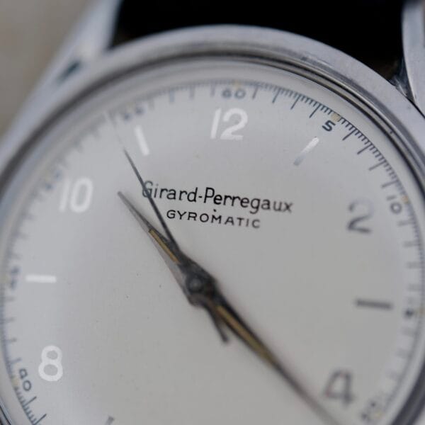 A close-up of the Girard Perregaux Gyromatic 6169 Caliber 47 watch face reveals silver-toned hour markers and hands on a white dial. The brand name and model are elegantly printed in black, all showcasing the time at approximately 10:09.