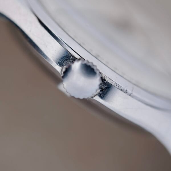 The Girard Perregaux Gyromatic 6169 Caliber 47 watch features a vintage charm with a metal crown displaying scratches and wear from time. Its shiny case has gentle signs of use, framed against a blurred neutral background that enhances its timeless elegance.