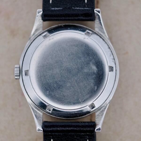 A close-up of the Girard Perregaux Gyromatic 6169 Caliber 47 shows its round metal casing with visible scratches, exuding charm. It has a black leather strap with white stitching, and the crown distinctively positioned on the left side.