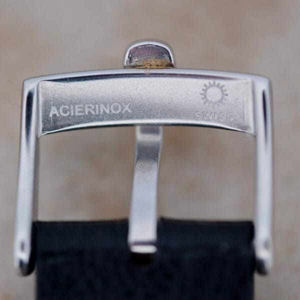 Close-up of the silver metal buckle from the Girard Perregaux Gyromatic 6169 Caliber 47, featuring ACIERINOX on the left, a sunburst logo next to SWISS on the right, and attached to a classic black strap. It exudes vintage charm.