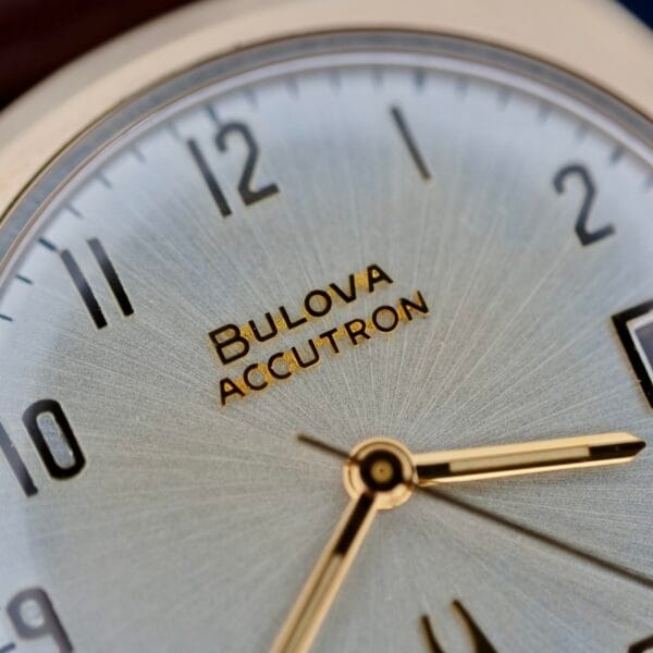 Close-up of a Bulova Accutron 218 Gold Tone watch. It features a silver dial with black Arabic numerals, gold hour and minute hands, and Bulova Accutron printed in gold below the center.