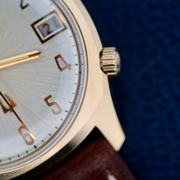 A close-up photo shows the Bulova Accutron 218 Gold Tone wristwatch, featuring a gold case and brown leather strap. The watch face displays numbers 3 to 6 with a date window at 3 oclock showing number 2, set against a dark blue background.