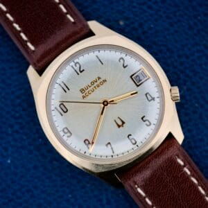 The Bulova Accutron 218 Gold Tone watch with a brown leather strap rests on a blue textured background, displaying the time at 2:10 and the date.