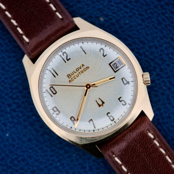 The Bulova Accutron 218 Gold Tone wristwatch features a gold-tone case with a brown leather strap. It has a silver dial with Arabic numerals, gold-tone hands, and a date window at 3 oclock, set against a textured blue background.