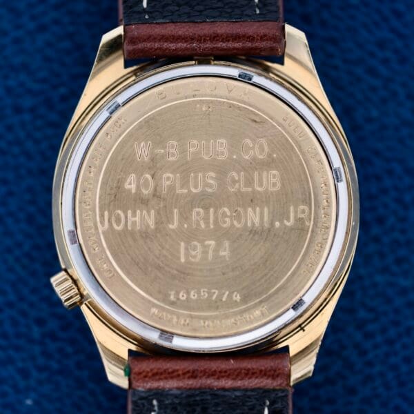 The image shows the back of a Bulova Accutron 218 Gold Tone wristwatch with a gold-toned case and black leather strap, engraved with W-B Pub. Co. 40 Plus Club John J. Rigoni, Jr. 1974. Its serial number is 1665714.