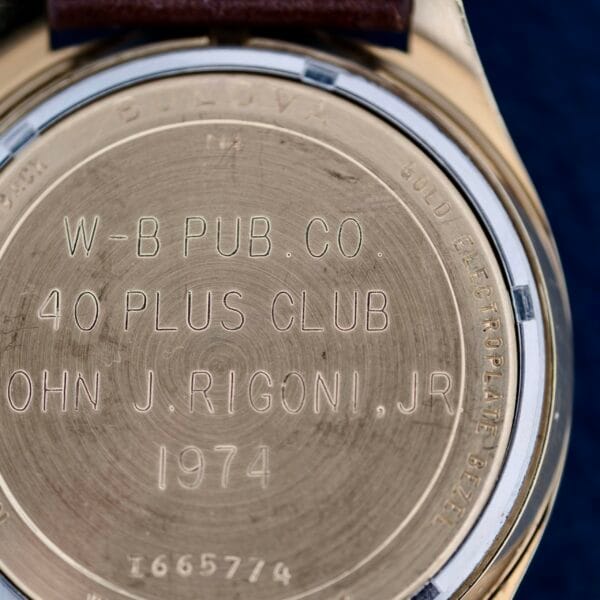 Close-up of a Bulova Accutron 218 Gold Tone watch back, featuring engraved text: W-B PUB. CO. 40 PLUS CLUB JOHN J. RIGONI, JR. 1974 and BULOVA GOLD ELECTROPLATE BEZEL, with serial number 1665774 visible at the bottom. The watch band is brown.