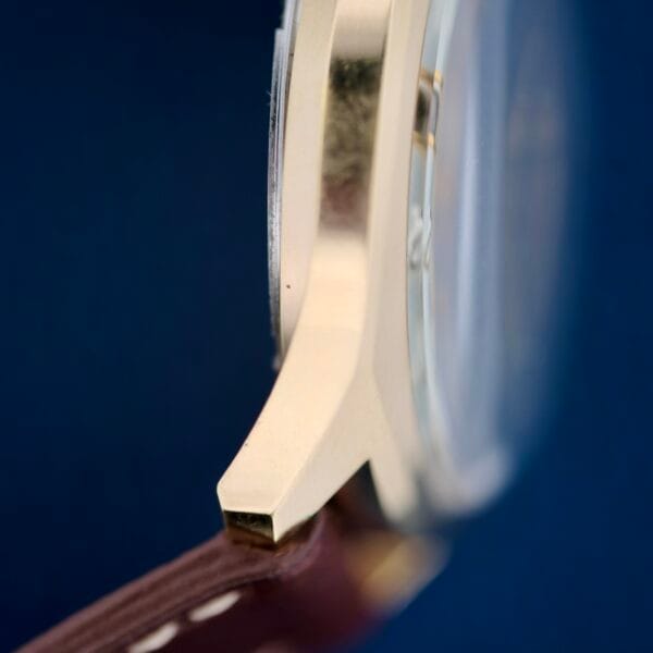 Close-up of a Bulova Accutron 218 Gold Tone wristwatch against a dark blue background, highlighting the metallic edge and clear glass face, with the brown leather strap and white stitching partially visible.