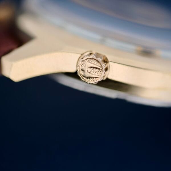 Close-up of the Bulova Accutron 218 Gold Tone watch crown with an engraved C, set against a dark blue background.