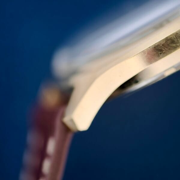 Close-up of the Bulova Accutron 218 Gold Tones side, showcasing its gold-colored case against a blurred blue backdrop. A hint of the brown leather strap is visible on the left, showing its attachment.