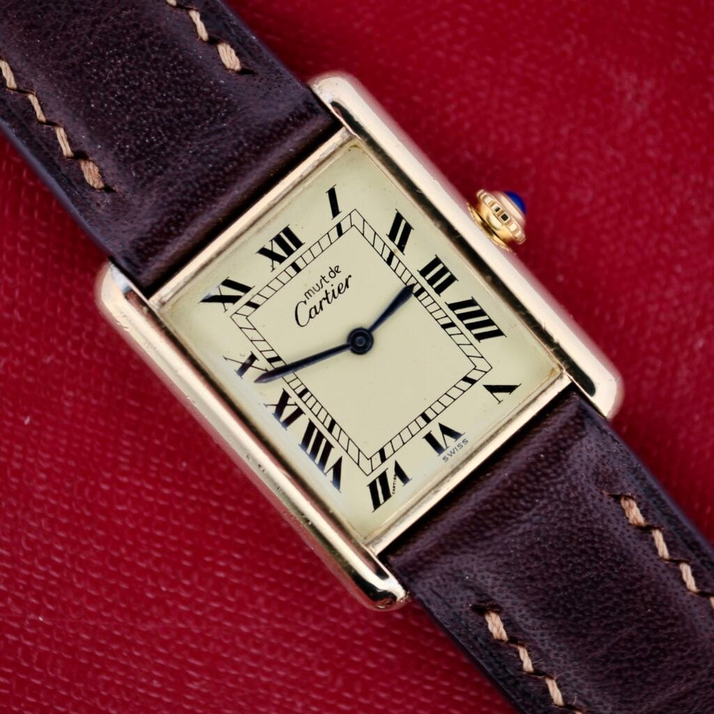 The Cartier Tank Must De Manual Wind, featuring a square gold case, Roman numerals, and a brown leather strap, rests on a red textured surface, embodying timeless elegance with its manual wind movement.