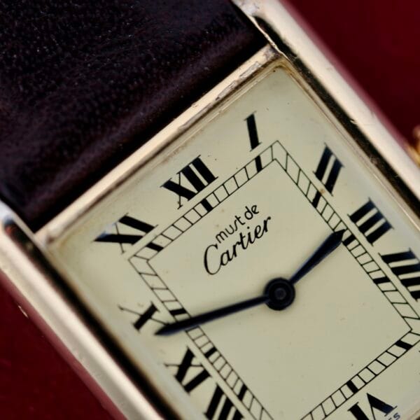 Close-up of a rectangular Cartier Tank Must De Manual Wind watch face, showing Roman numerals and black hands, with must de Cartier text. The gold-tone case and dark leather strap are set against a blurred red background.