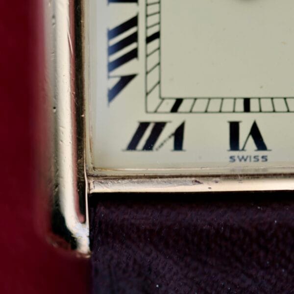 Close-up of the Cartier Tank Must De Manual Wind watch face displaying Roman numerals VII, VIII, IX; and SWISS. It features a gold-colored bezel with a partially visible dark red frame.