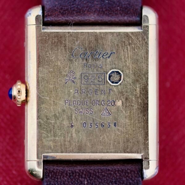 A close-up of the Cartier Tank Must De Manual Wind watch reveals an engraved rose symbol, 925 ARGENT, PLAQUE OR G 20M SWISS, and numbers 6 03534. It features a brown leather strap and a crown adorned with a blue gem.