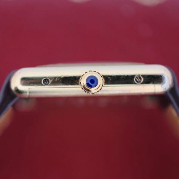 Close-up of the Cartier Tank Must De Manual Winds gold case crown, featuring a blue cabochon. The watch strap is visible on both sides against a blurred red background.