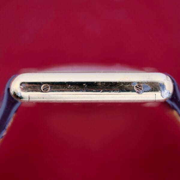 A close-up of the Cartier Tank Must De Manual Wind from the side reveals a gold-colored frame with two visible screws. The watch has a dark band and is set against a solid red background.
