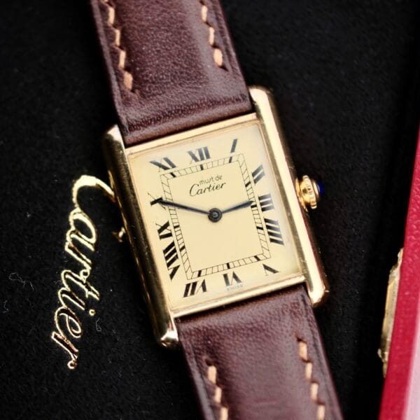 The Cartier Tank Must De Manual Wind, a rectangular gold wristwatch with a brown leather strap, is set against a textured black surface. It has a cream dial with Roman numerals and blue-tinted hands.