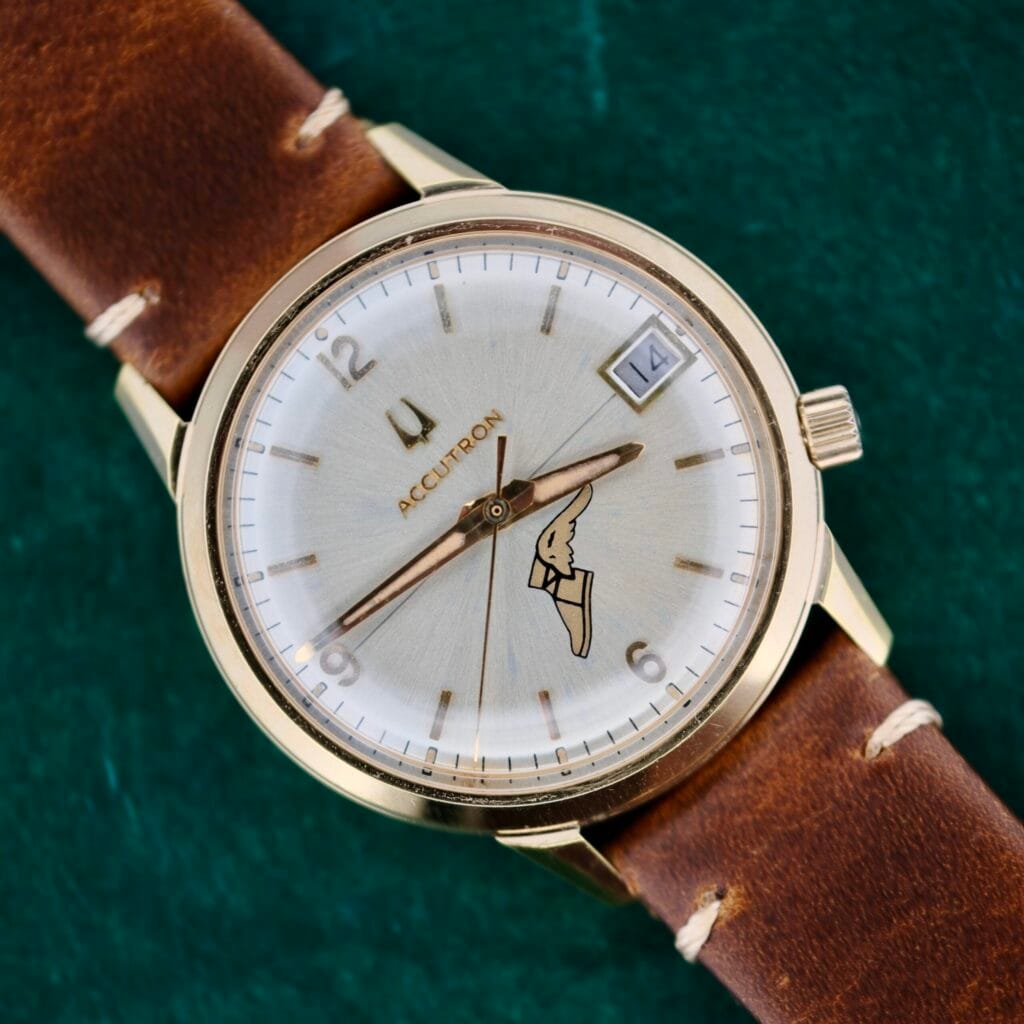 Bulova Accutron 218 Goodyear Dial wristwatch featuring a gold case, brown leather band, date display, and distinctive hands, shown on a green background.