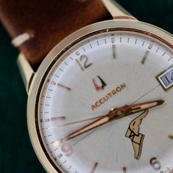 A close-up of a vintage Bulova Accutron 218 Goodyear wristwatch features a cream dial with silver hour markings, a winged logo above the 6 oclock position, the date window displaying 14, and is complemented by a brown leather strap and gold-toned case.