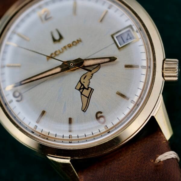 A close-up of the vintage Bulova Accutron 218 Goodyear Dial wristwatch highlights its round gold case, classic white dial with gold hour markers and hands, a date window at 3 oclock, and distinctive logo at the center, all complemented by a brown leather strap for timeless appeal.