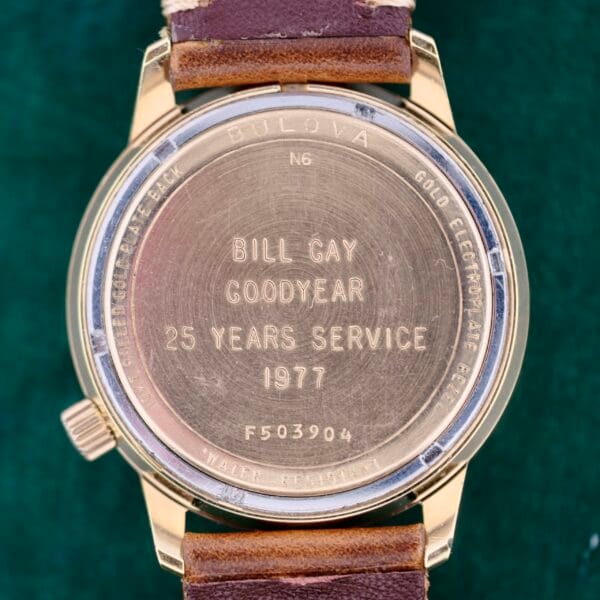 Close-up of a gold Bulova Accutron 218 Goodyear Dial watch back, engraved with Bill Gay Goodyear 25 Years Service 1977. Surrounding text reads Water Resistant and Gold Electroplate Bezel. Partially visible brown leather straps are included.