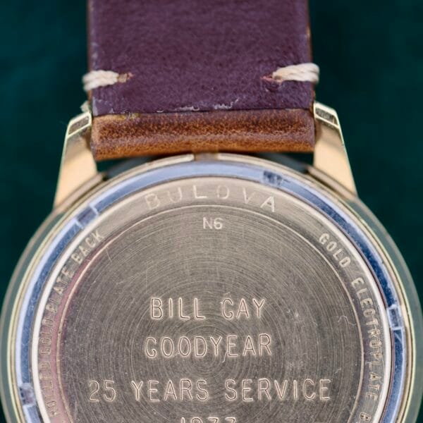 The image displays the back of a Bulova Accutron 218 Goodyear Dial, engraved with BILL GAY GOODYEAR 25 YEARS SERVICE 1973. Its hidden face contrasts with a brown leather strap featuring visible stitching, offering timeless appeal.