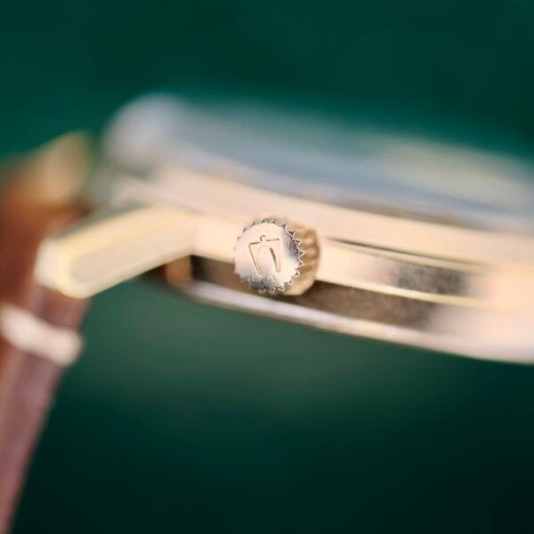 Close-up of the Bulova Accutron 218 Goodyear Dial, highlighting its crown with a small etched logo. The watch features a gold case and brown leather strap, set against a blurred dark green background.
