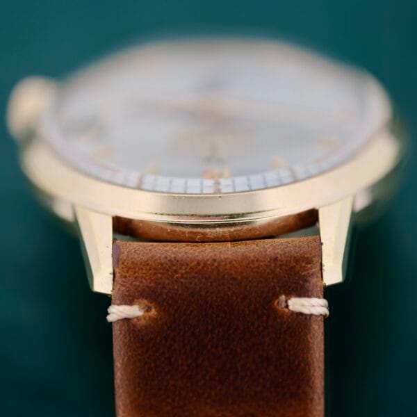 A close-up shows a Bulova Accutron 218 Goodyear Dial with a brown leather strap gently embracing its gold-toned casing. The face is out of focus, obscuring details, against a muted green background that exudes timeless elegance.