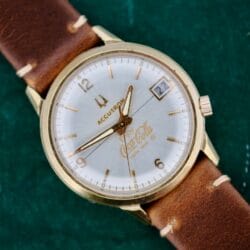 The Bulova Accutron 218 Coca-Cola Dial is a vintage wristwatch with a brown leather strap, gold bezel, white face, gold hour markers and hands. It features a date display at 3 oclock with Coca-Cola and Time King printed on the dial.