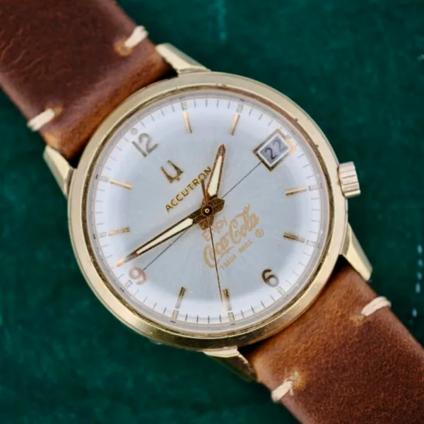 The Bulova Accutron 218 Coca-Cola Dial is a vintage wristwatch with a brown leather strap, gold bezel, white face, gold hour markers and hands. It features a date display at 3 oclock with Coca-Cola and Time King printed on the dial.