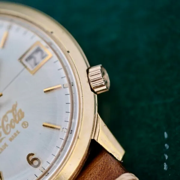Close-up of a Bulova Accutron 218 Coca-Cola dial wristwatch with a gold case, brown leather strap, and gold hour markers. A date window sits at the 3 oclock position against a dark green background.