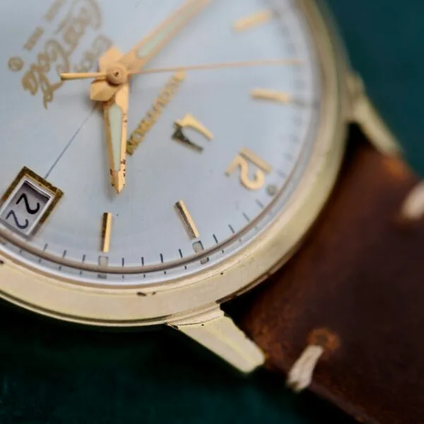 A close-up of the Bulova Accutron 218 Coca-Cola Dial wristwatch showcases a round, gold-colored case with a brown leather strap. It features gold hour markers, a date display showing the 22nd, and its hands pointing at 12 and 4.