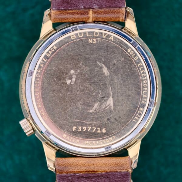 The image displays the back of a gold-colored Bulova Accutron 218 Coca-Cola Dial watch, featuring a side crown, engraved text including Bulova, Gold Electroplate Bezel, and Water Resistant, along with the serial number F397716. It also has a brown leather strap.