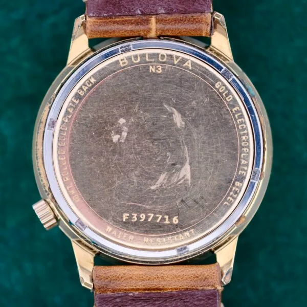 The image displays the back of a gold-colored Bulova Accutron 218 Coca-Cola Dial watch, featuring a side crown, engraved text including Bulova, Gold Electroplate Bezel, and Water Resistant, along with the serial number F397716. It also has a brown leather strap.