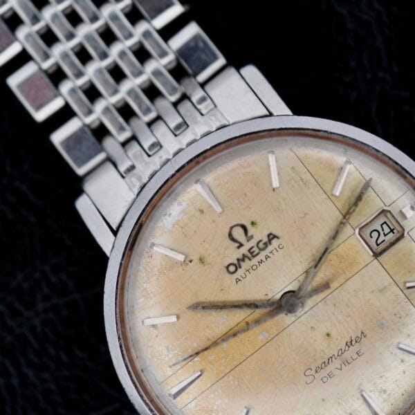 Close-up of an Omega Seamaster Deville Automatic 166.020 watch, featuring a vintage-style yellowed dial, silver hour markers, and a date window at 3 oclock. The metal bracelet is partially visible against a dark background.