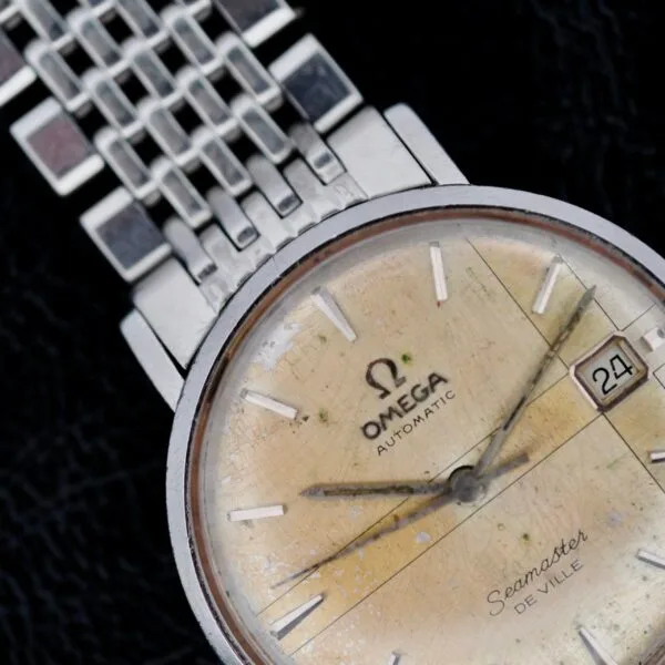 Close-up of an Omega Seamaster Deville Automatic 166.020 watch, featuring a vintage-style yellowed dial, silver hour markers, and a date window at 3 oclock. The metal bracelet is partially visible against a dark background.