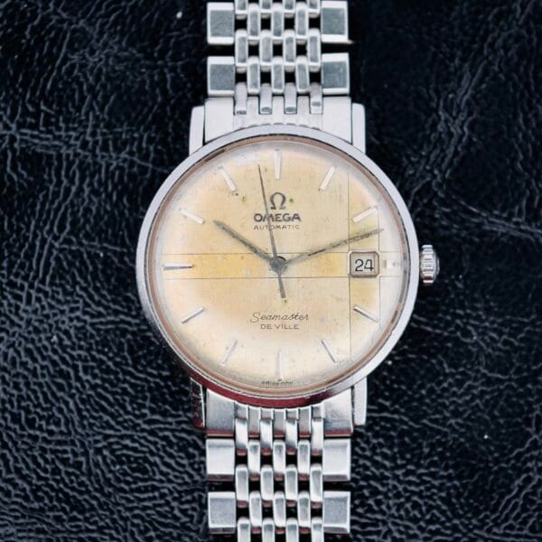 A vintage Omega Seamaster Deville Automatic 166.020 wristwatch features a silver mesh bracelet, beige dial, and textured black leather background. The dial includes the Omega logo, Automatic, Seamaster De Ville, and a date window showing 24.