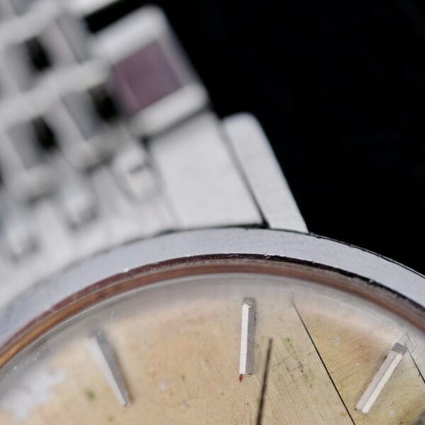 The image features a close-up of an Omega Seamaster Deville Automatic 166.020 wristwatch with a metal band. It showcases the beige dial with rectangular hour markers and partially visible square metal link band in the background, slightly out of focus.